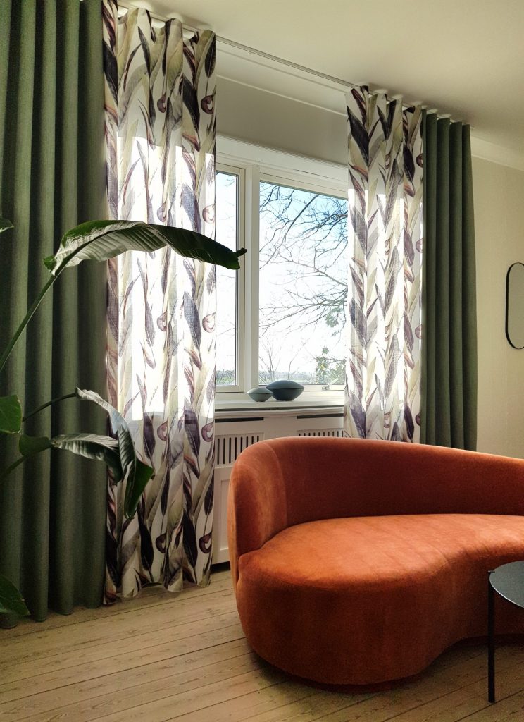 Modern eingerichtetes Wohnzimmer mit grünen Dekoschals und transparenten Stores mit Blattmuster von Pagunette. Ergänzt durch ein orangenes Sofa, Holzdielenboden und schwarze Akzente