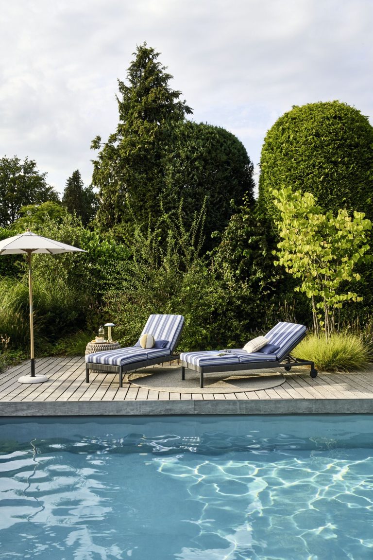 Zwei gepolsterte Sonnenliegen mit gestreiften Outdoor Stoffen auf einer Holzterrasse am Pool, daneben ein Sonnenschirm und bepflanzter Garten