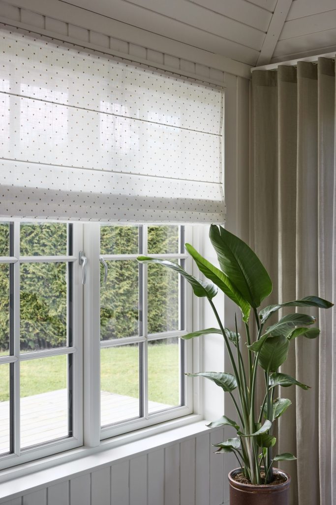 Helles Fenster mit halbtransparentem Raffrollo mit feinem Punktmuster und seitlichem Vorhang in Naturton, davor eine große grüne Zimmerpflanze