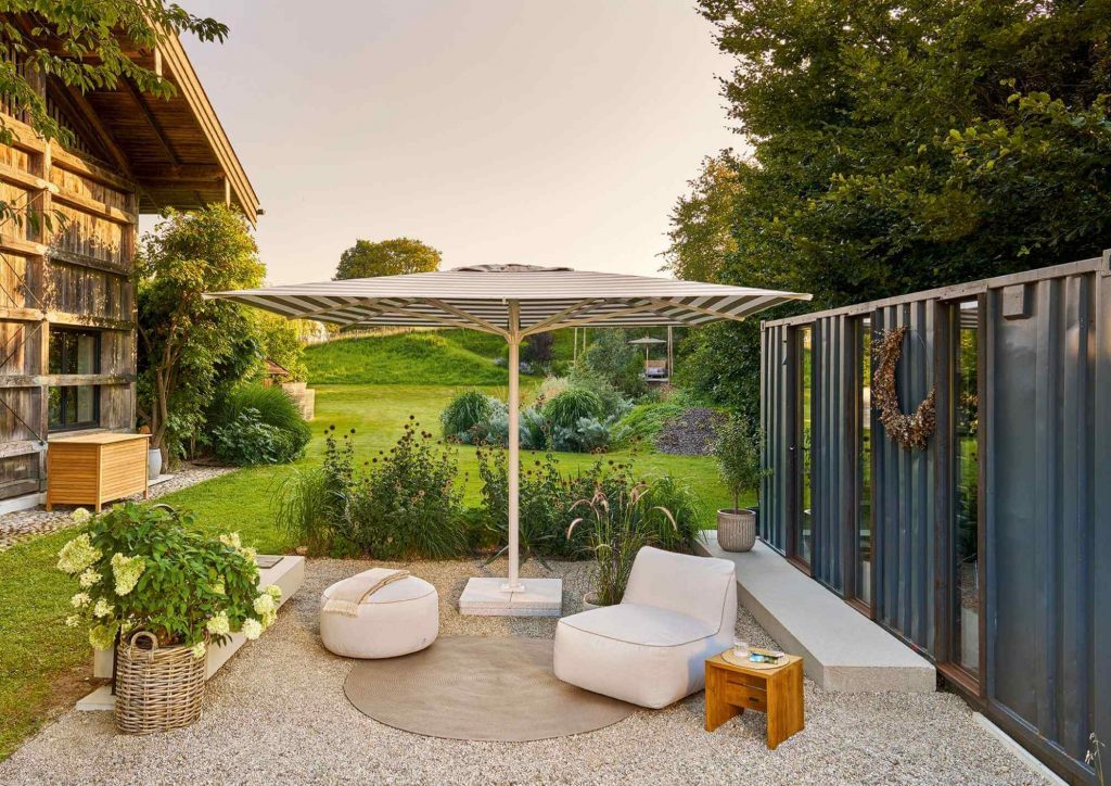 Großer gestreifter Weishäupl Sonnenschirm auf einem Kiesgarten mit weißem Lounge-Sessel und Blick ins Grüne