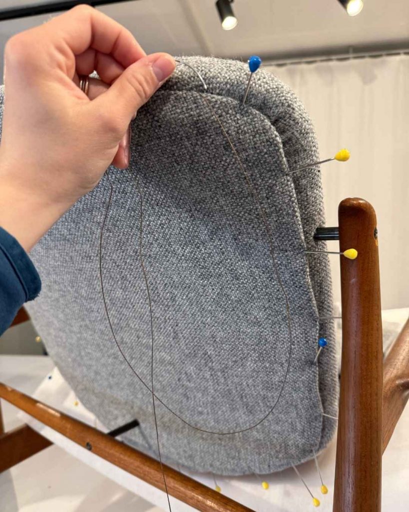 Hand beim Aufziehen des hellgrauen Wollstoffs über die Rückenlehne eines Finn Juhl 136 mit Polsterstecker zur Fixierung