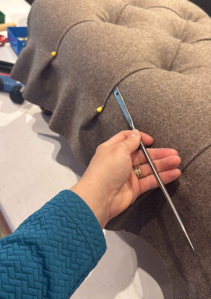 Hand mit einer langen Polsternadel an einem braun bezogenen Fußschemel während der handwerklichen Ausarbeitung der Rautenheftung.