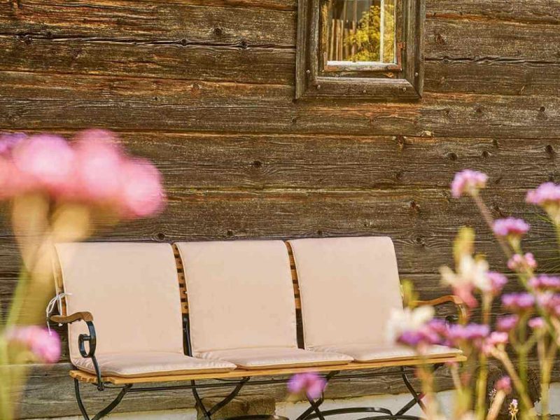 Weishäupl Gartenbank mit cremefarbenen Sitz- und Rückenpolstern vor rustikaler Holzfassade mit Wildblumen