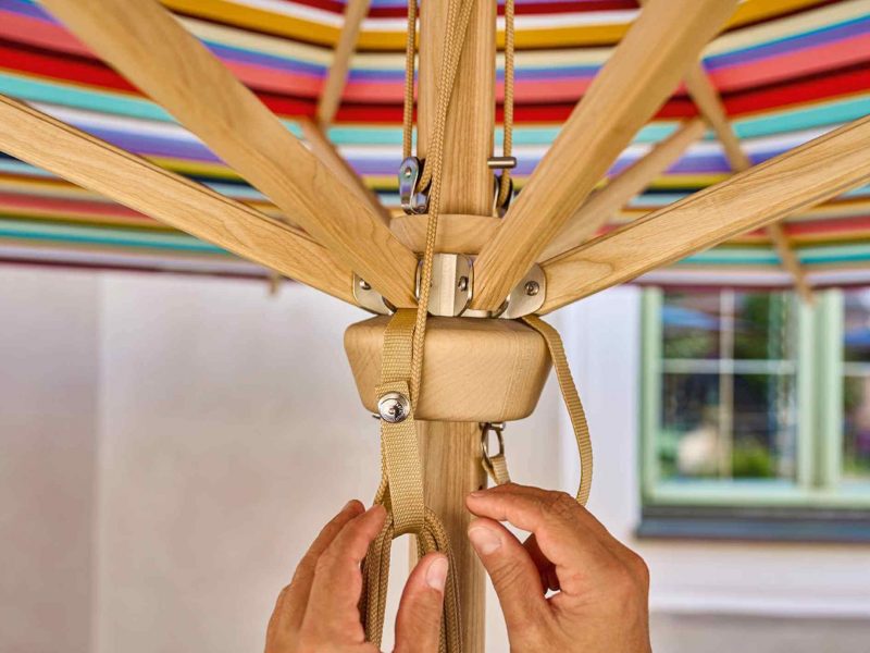 Hände bedienen den Holzmechanismus eines Weishäupl Sonnenschirms mit buntem Multicolor-Bezug