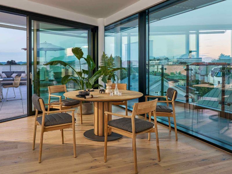 Runder Weishäupl Teva Tisch mit Teakholzstühlen und Polstern vor einer großen Glasfront mit Blick auf die Terrasse