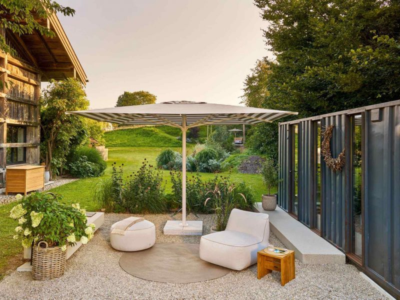 Großer gestreifter Weishäupl Sonnenschirm auf einem Kiesgarten mit weißem Lounge-Sessel und Blick ins Grüne