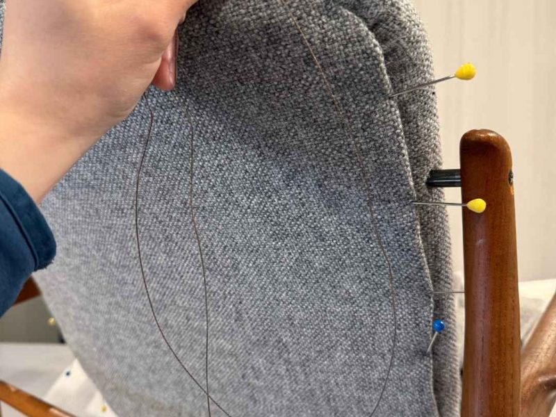 Hand beim Aufziehen des hellgrauen Wollstoffs über die Rückenlehne eines Finn Juhl 136 mit Polsterstecker zur Fixierung