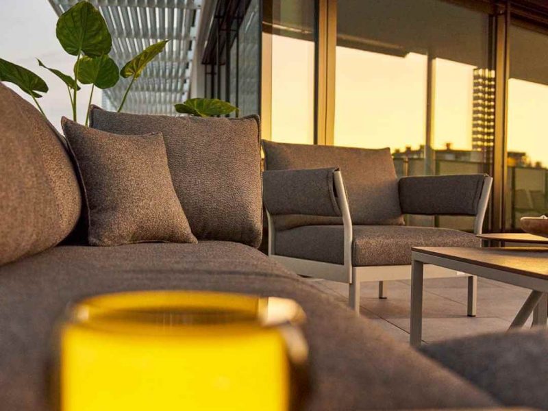 Weishäupl Loungesofa mit grauen Polstern auf einer Dachterrasse im Sonnenuntergang, im Vordergrund eine leuchtende Outdoorlampe