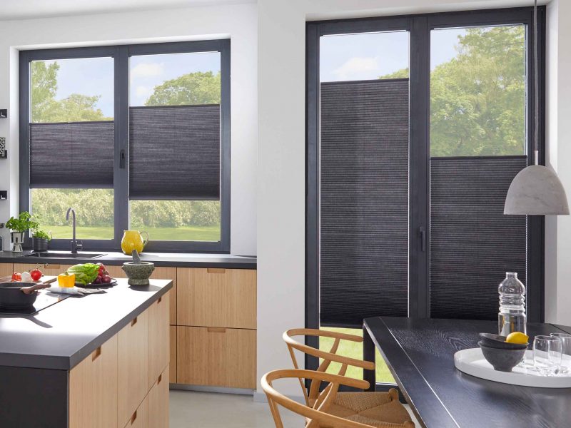 Moderne Küche mit dunklen Fensterrahmen und schwarzen Plissees zur Verdunkelung, Holztisch mit Stühlen im Vordergrund