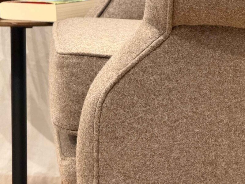 Nahaufnahme eines neu bezogenen Ohrensessels mit braunem Schurwollstoff, sauber verarbeitetem Keder und geschwungenem Holzfuß.