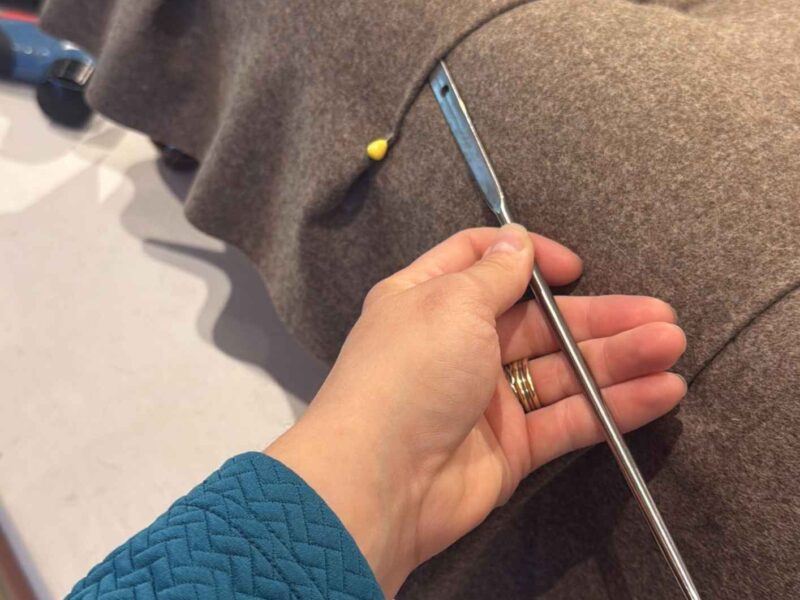 Hand mit einer langen Polsternadel an einem braun bezogenen Fußschemel während der handwerklichen Ausarbeitung der Rautenheftung.